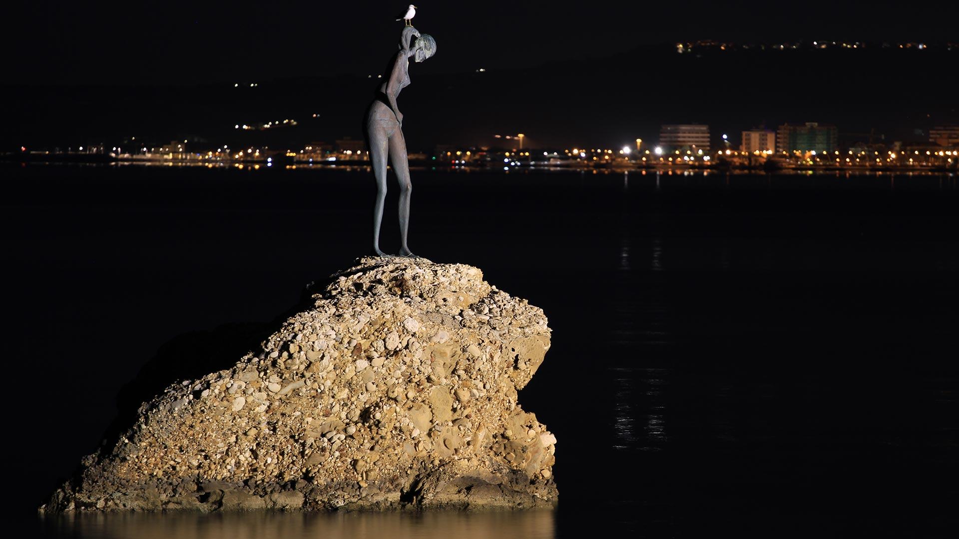 Statua su una roccia illuminata di notte, città sullo sfondo.
