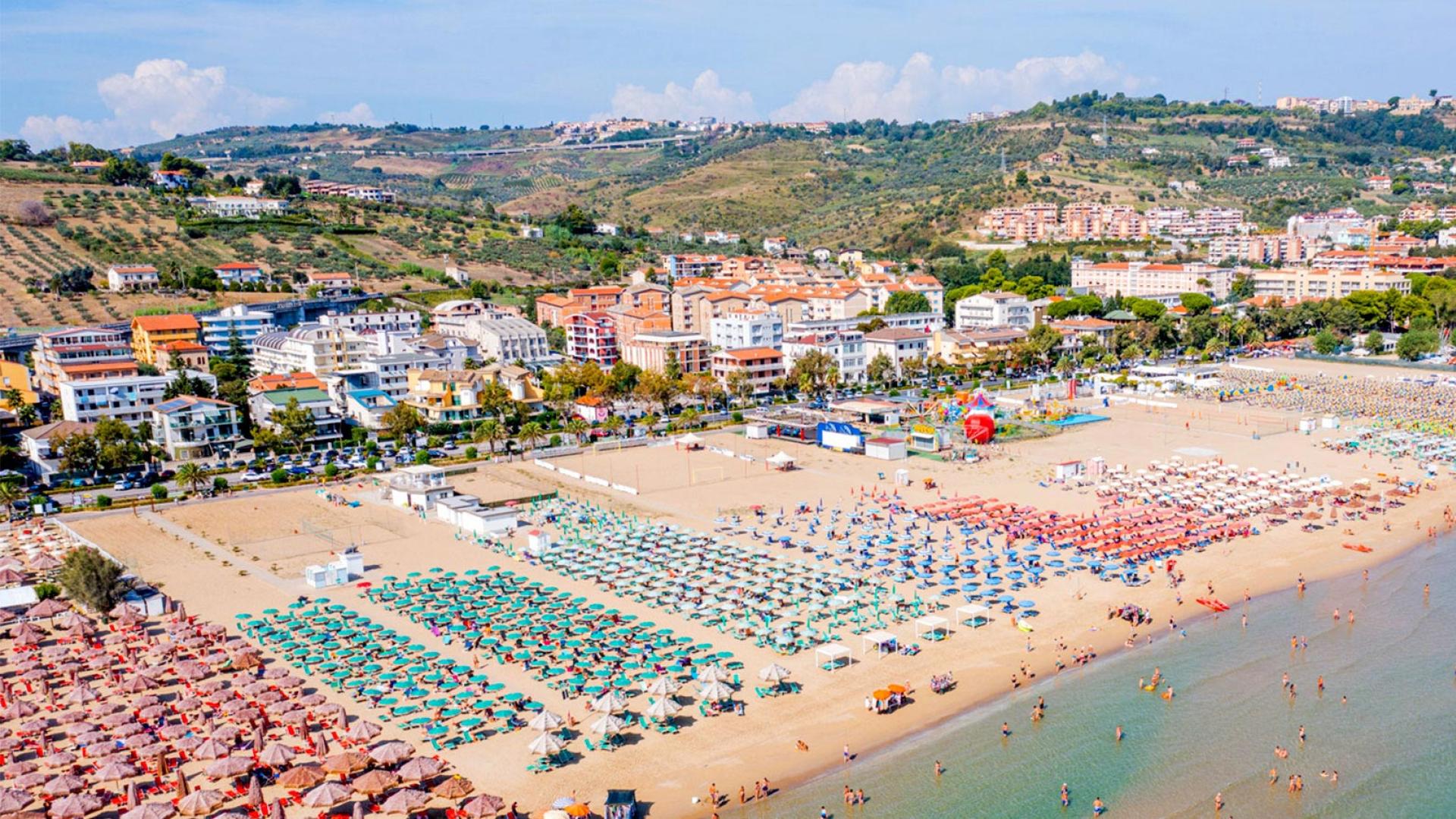Spiaggia affollata con ombrelloni colorati, edifici e colline sullo sfondo.