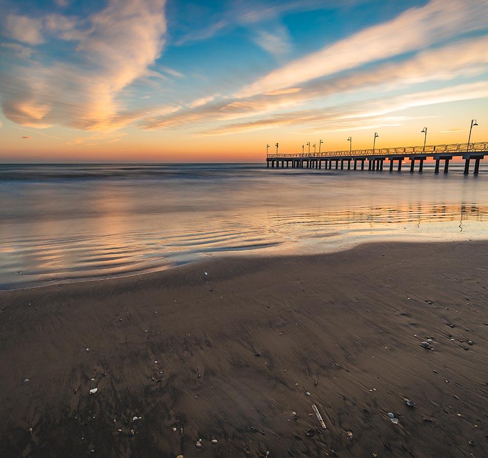 Sunset on a beach with a long pier and cloudy sky.
