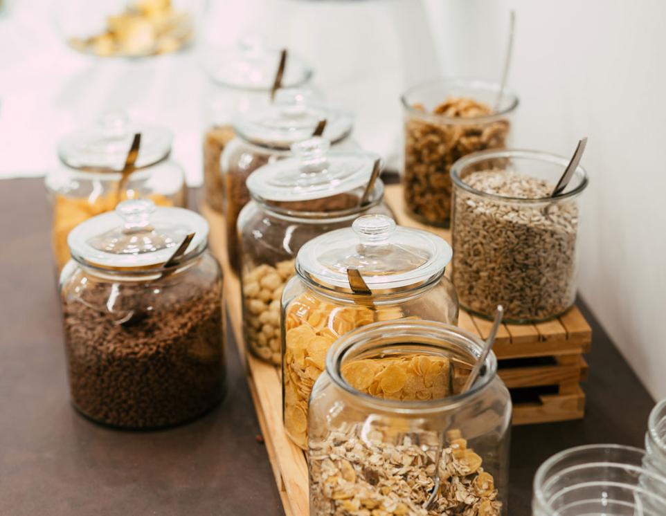 Barattoli di cereali e semi su un tavolo, con bicchieri di vetro accanto.